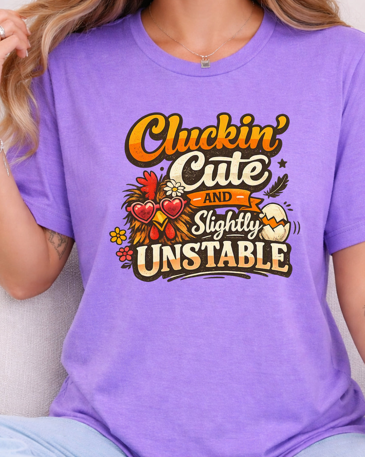 A woman wearing a purple graphic t-shirt with the text 'Cluckin' Cute and Slightly Unstable' in a retro font, decorated with autumn-themed illustrations including hearts, leaves, and a cracked egg.