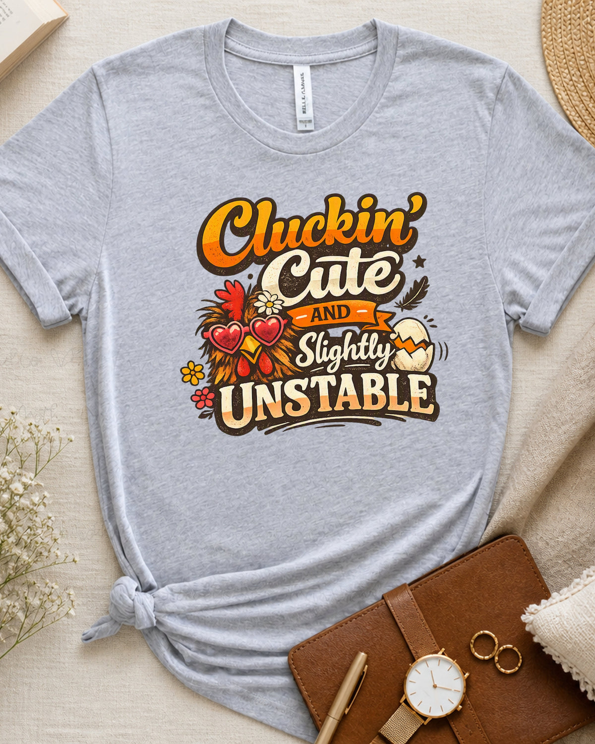 A heather gray short-sleeve t-shirt with a graphic that reads 'Cluckin' Cute and Slightly Unstable' in orange, yellow, and brown lettering, styled in a flat lay with a brown leather notebook, a watch, rings, and dried flowers on a beige fabric background.