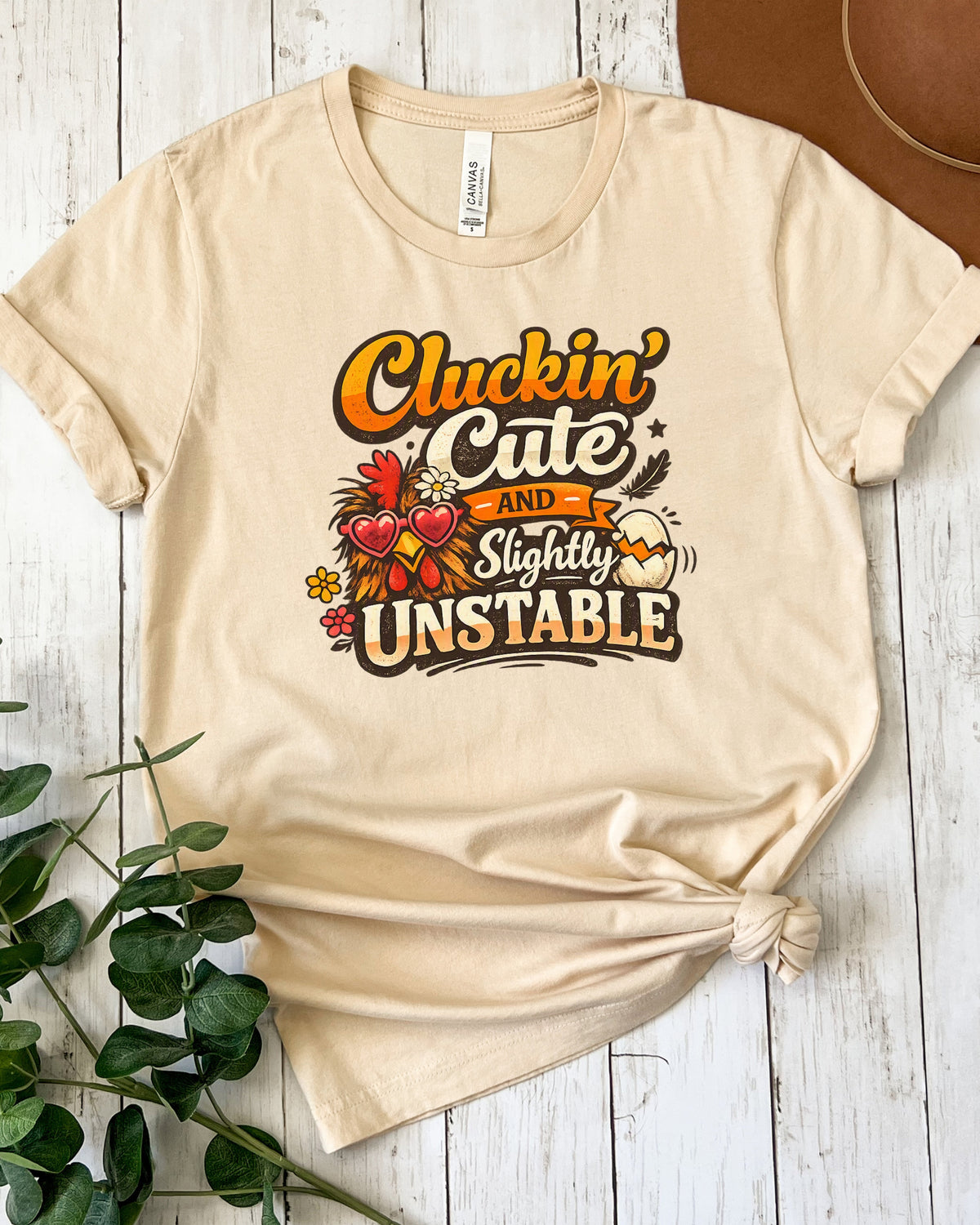 A beige graphic t-shirt with rolled sleeves, styled in a flat-lay on a white wood background with a brown hat and green leaves. The shirt features a graphic that reads 'Cluckin' Cate and Slightly UNSTABLE' with a cartoon turkey.
