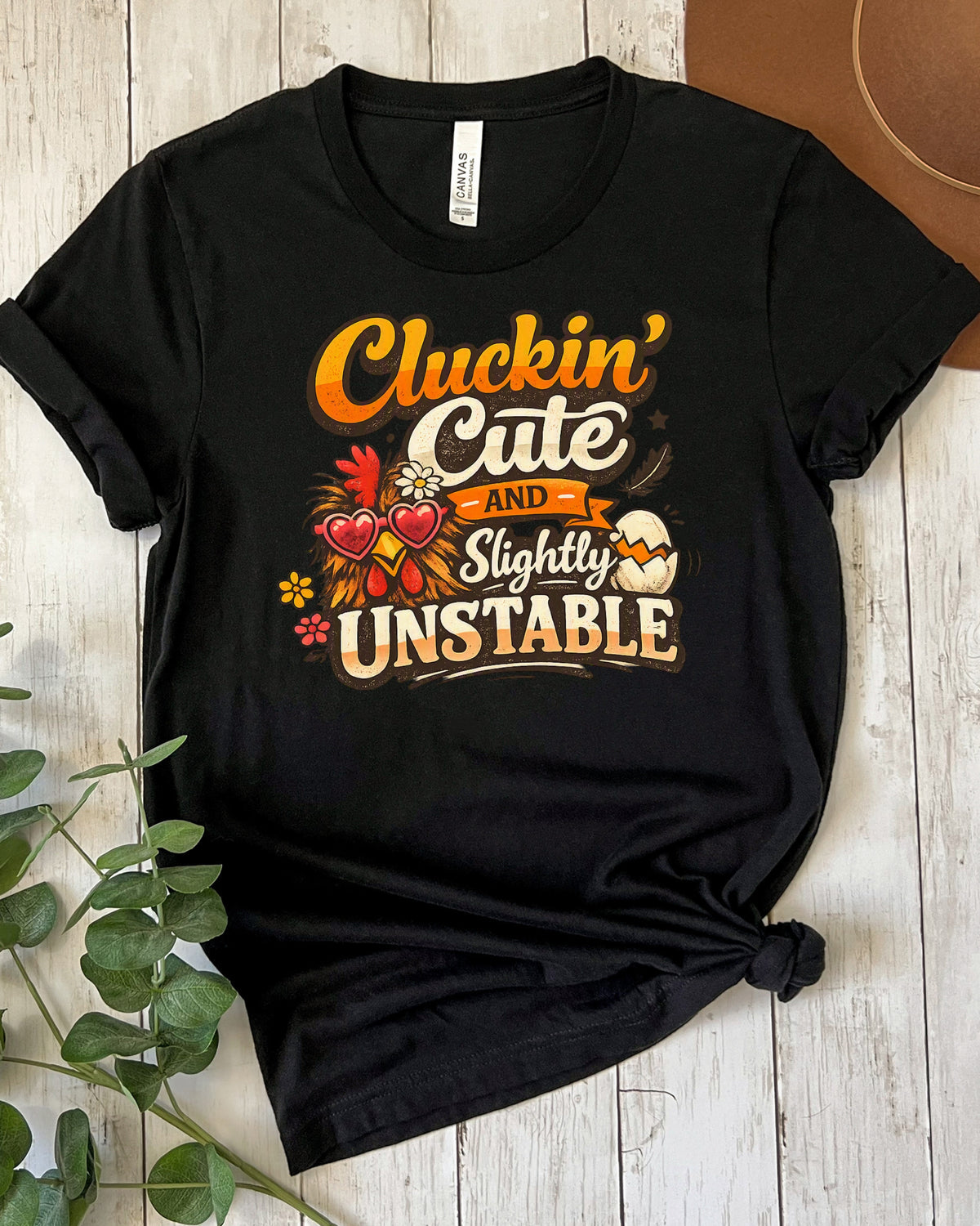 A black graphic t-shirt featuring a humorous autumn-themed design that reads 'Cluckin' Cute and Slightly Unstable' with a rooster, hearts, and an Easter egg, styled on a white wood plank background.