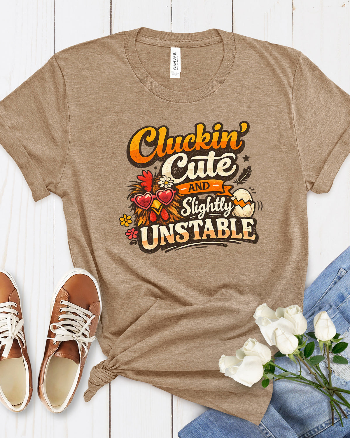 A heather tan graphic t-shirt laid flat on a white wood background, styled with brown sneakers, white roses, and blue jeans. The t-shirt features a graphic that reads 'Cluckin' Cute and Slightly Unstable' in orange, yellow, and brown lettering with red hearts and autumn-themed illustrations.