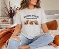 A woman sitting cross-legged on a bed wearing a light heather gray graphic t-shirt with the text 'WALKING BY FAITH tripping constantly' and three cartoon dogs tripping. The setting is a cozy bedroom with autumn decor, including a knit blanket, pumpkins, and a sign.