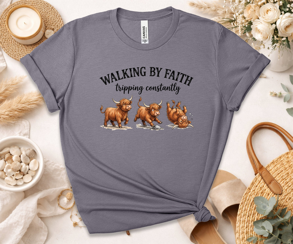 A heather gray graphic t-shirt featuring three cartoon Highland cows tripping, with the text 'WALKING BY FAITH tripping constantly'. The t-shirt is styled in a flat-lay arrangement with a candle, dried grass, a straw hat, and a woven bag on a white background.