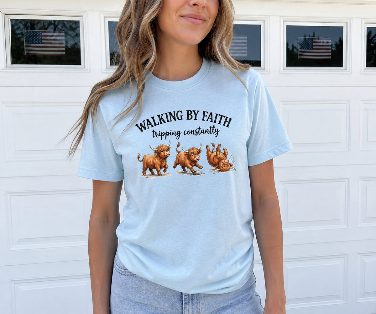 A woman wearing a light blue graphic t-shirt with the text 'WALKING BY FAITH tripping constantly' and illustrations of three brown dogs, standing in front of a white garage door.