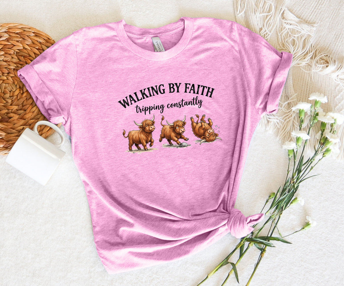 A pink short-sleeve graphic T-shirt laid flat on a white textured surface with decorative props including a woven mat, white flowers, and a small white mug. The shirt has black text reading 'WALKING BY FAITH tripping constantly' above three cartoon pigs walking.
