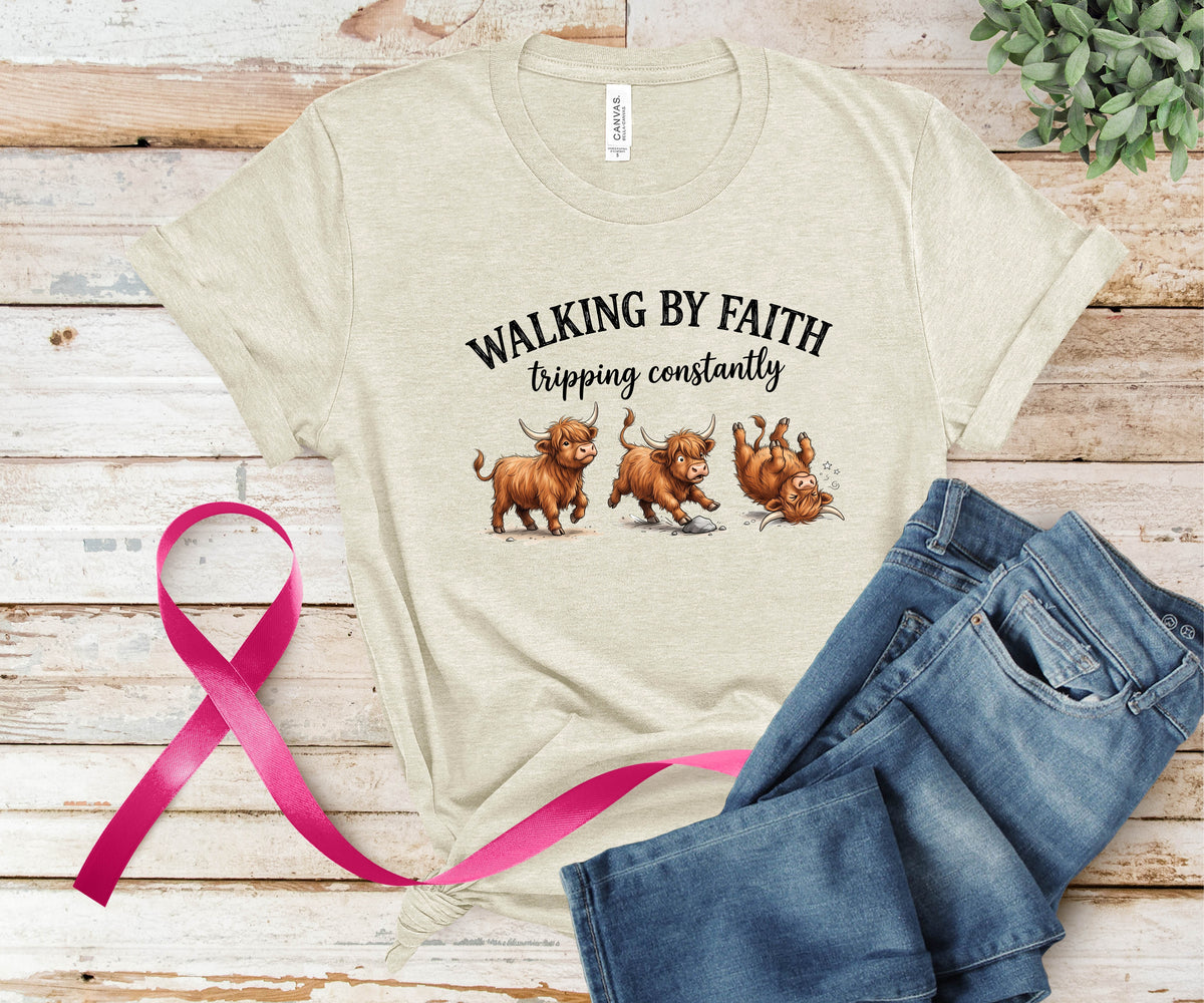 A beige graphic t-shirt featuring three Highland cattle illustrations and the text 'WALKING BY FAITH tripping constantly', styled in a flat lay on a rustic wooden background with blue jeans and a pink ribbon.