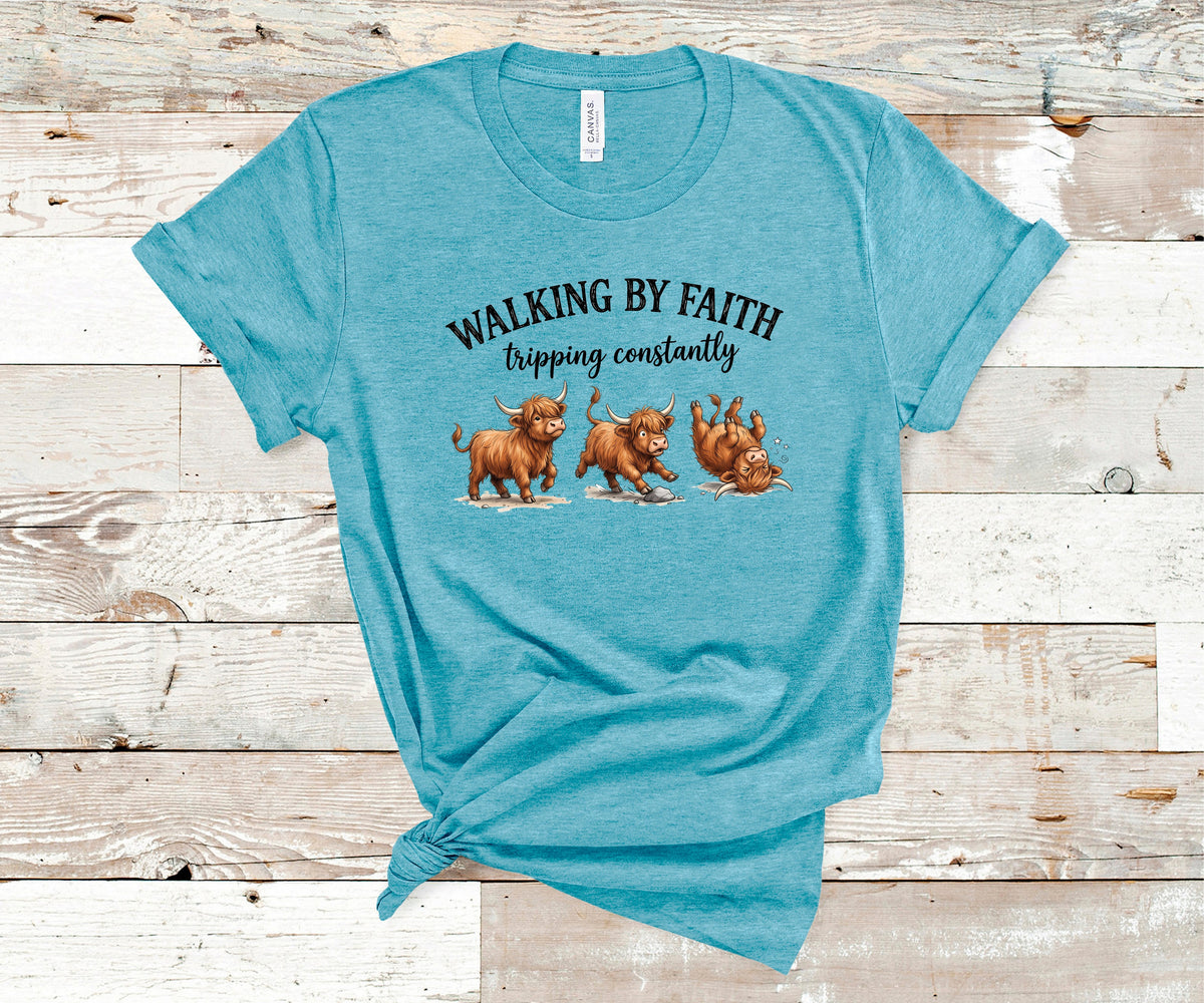 A heather teal t-shirt with a graphic print of three brown dogs and the text 'WALKING BY FAITH tripping constantly', laid flat on a rustic white-washed wood plank background.