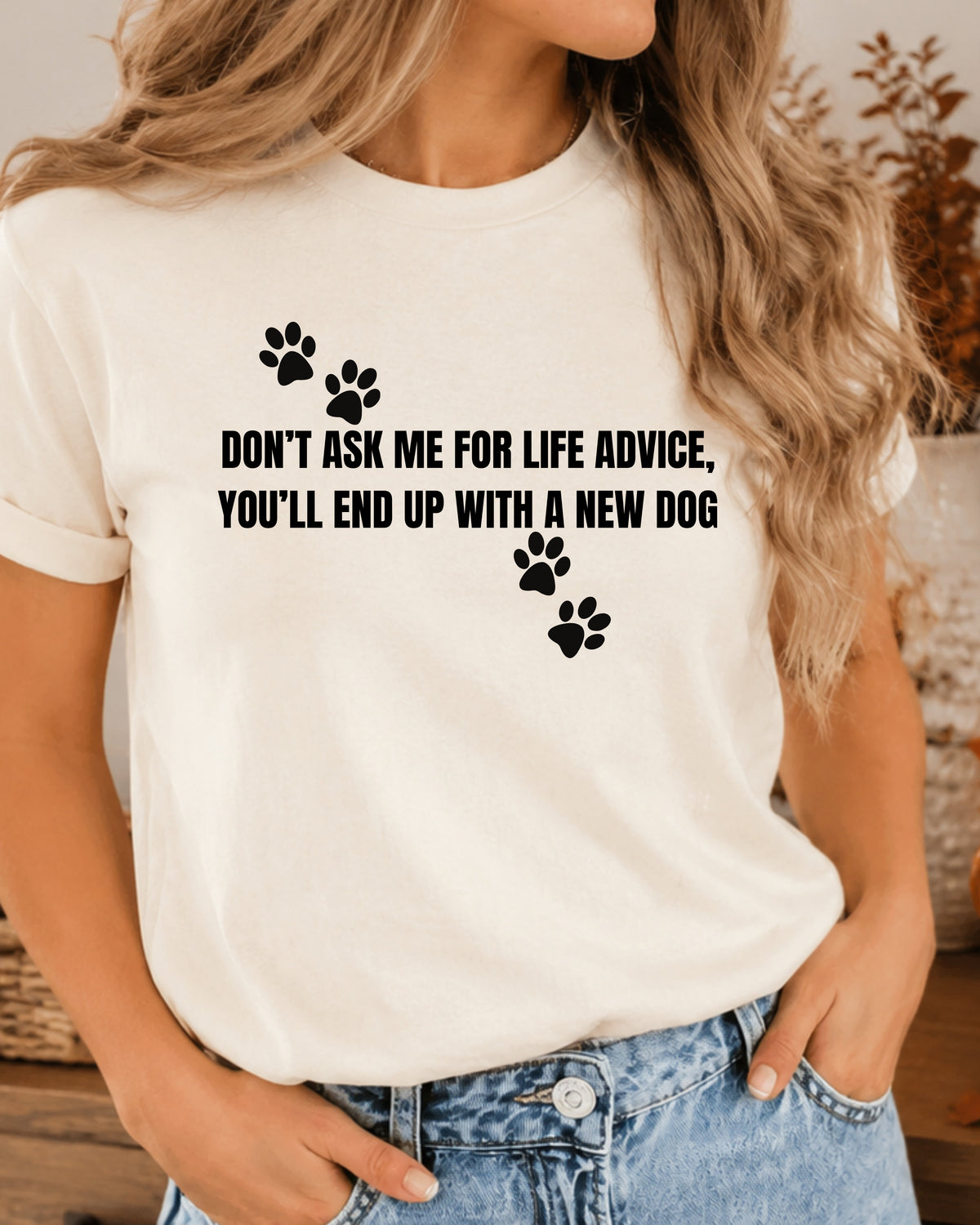 A woman wearing a white short-sleeve graphic t-shirt with black paw prints and the text 'DON'T ASK ME FOR LIFE ADVICE, YOU'LL END UP WITH A NEW DOG', paired with blue jeans.