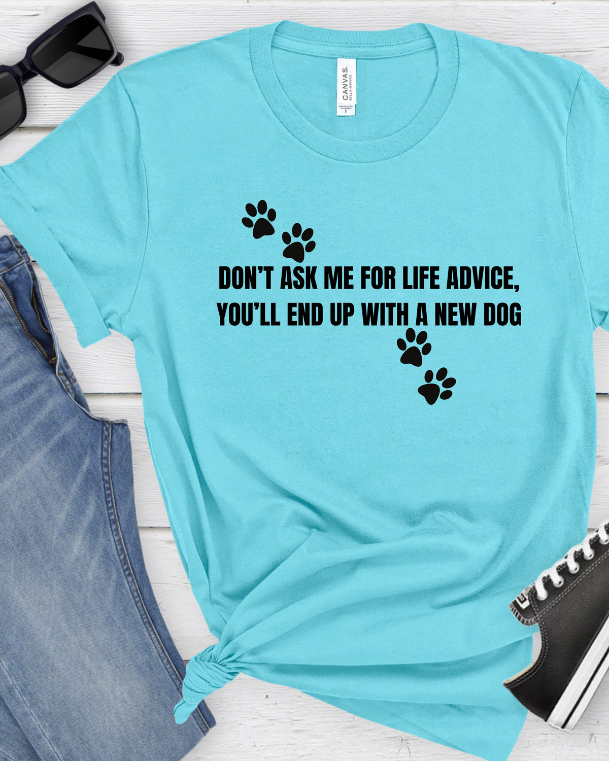 A light blue t-shirt with black text reading 'DON'T ASK ME FOR LIFE ADVICE, YOU'LL END UP WITH A NEW DOG' and black paw prints, styled in a flat lay with jeans, sunglasses, and a shoe on a white wood background.