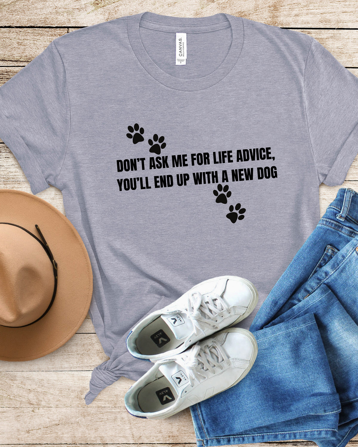 A heather gray graphic t-shirt with black text and paw prints, styled in a flat lay with a tan hat, blue jeans, and white sneakers on a light wood background.