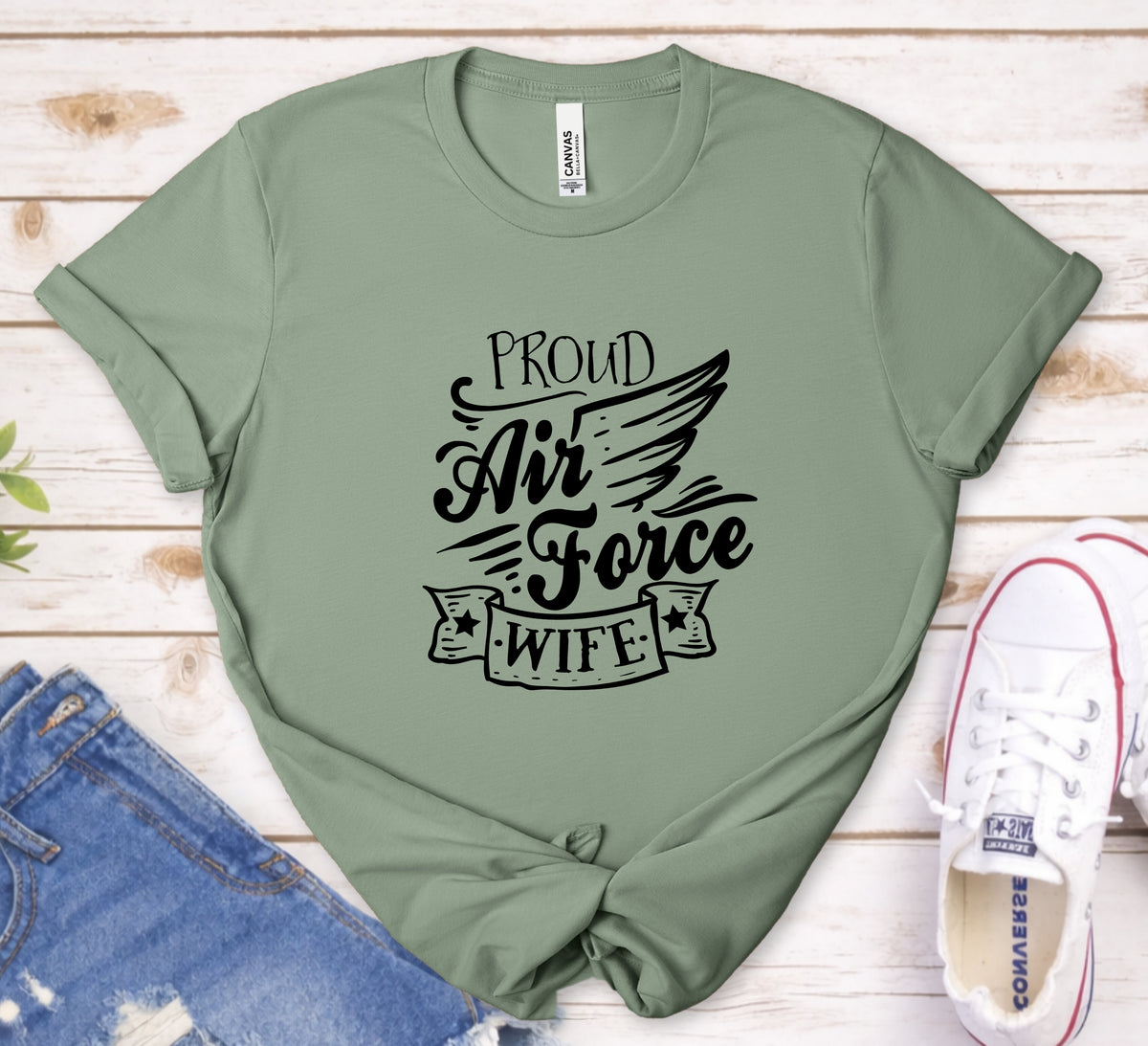 An olive green short-sleeve t-shirt with a black graphic reading 'PROUD Air Force WIFE' and a wing design, styled in a flat lay on a white wood background with blue jeans and white Converse sneakers.