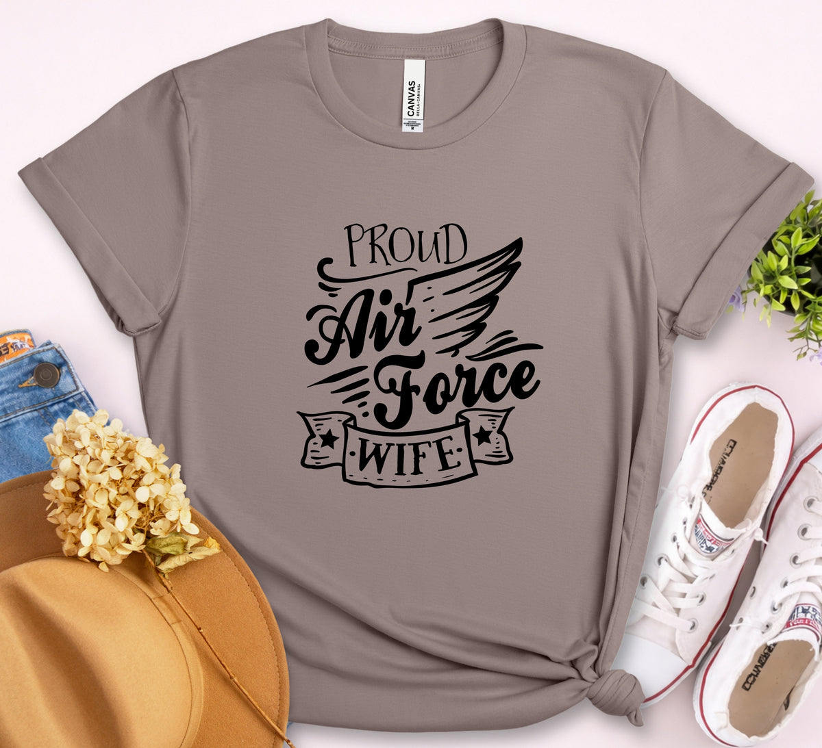 A taupe gray short-sleeve t-shirt with a black graphic reading 'PROUD Air Force WIFE' styled in a flat-lay with a tan hat, blue jeans, white sneakers, and dried flowers on a light pink background.