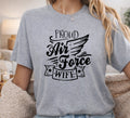 A woman wearing a heather gray graphic t-shirt with the text 'PROUD Air Force WIFE' in black script and wing design, photographed in a cozy indoor setting.