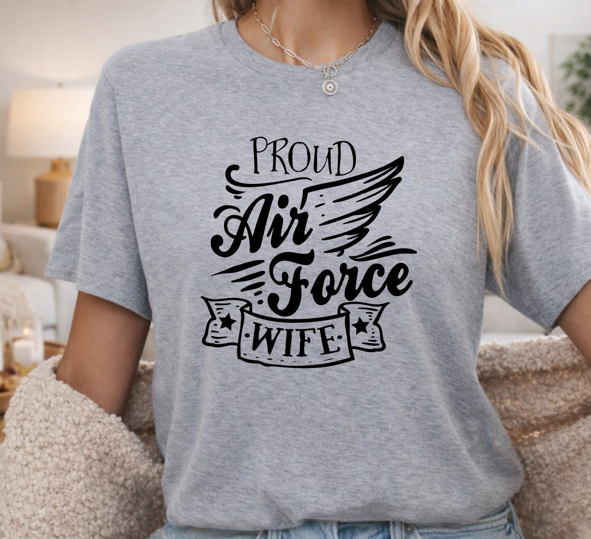 A woman wearing a heather gray graphic t-shirt with the text 'PROUD Air Force WIFE' in black script and wing design, photographed in a cozy indoor setting.