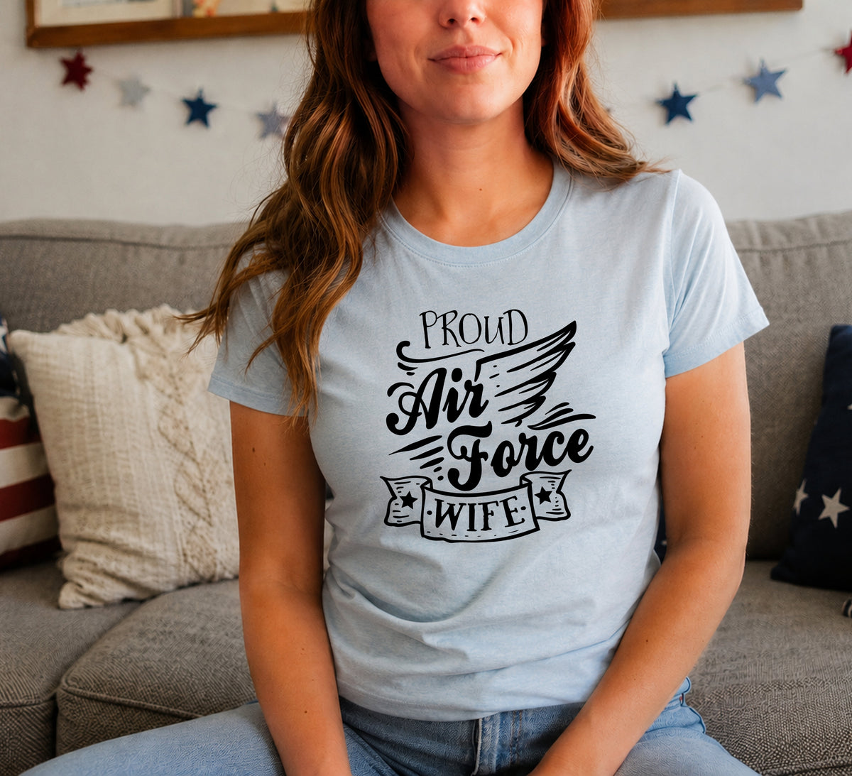 A woman wearing a light blue crewneck t-shirt with a black graphic that reads 'PROUD Air Force WIFE' sitting on a gray couch in a living room.