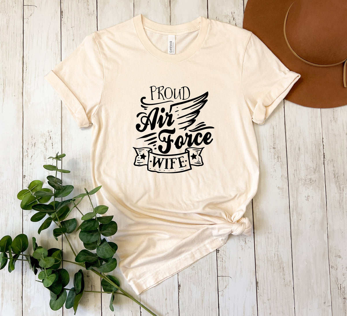 A cream-colored short-sleeve graphic t-shirt with the text 'PROUD Air Force WIFE' in black script, styled in a flat lay on a white wood plank background with a brown hat and green eucalyptus leaves.