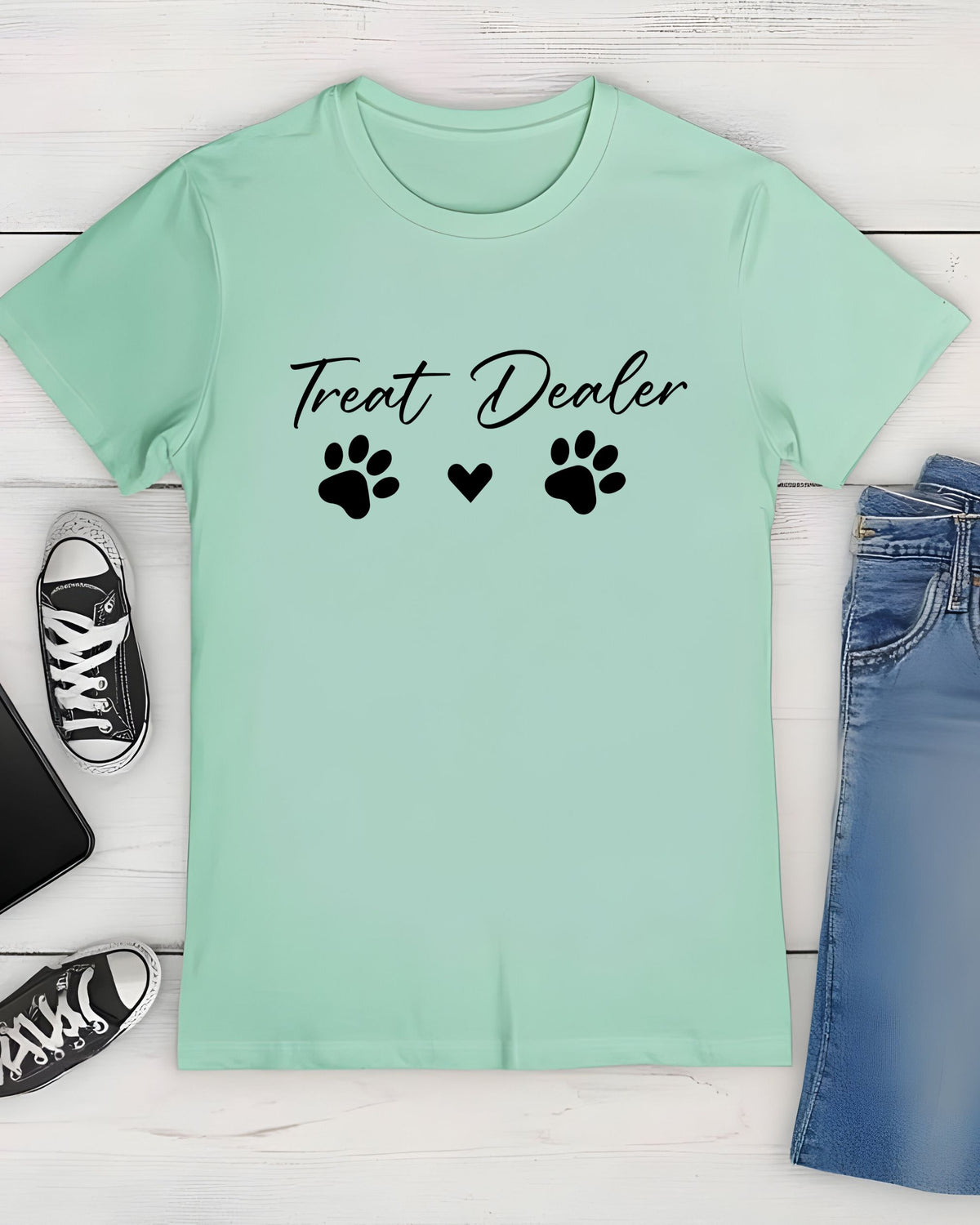 A mint green short-sleeve t-shirt with 'Treat Dealer' and paw prints printed in black on the chest, styled in a flat lay with jeans, sneakers, and a phone on a white wood background.