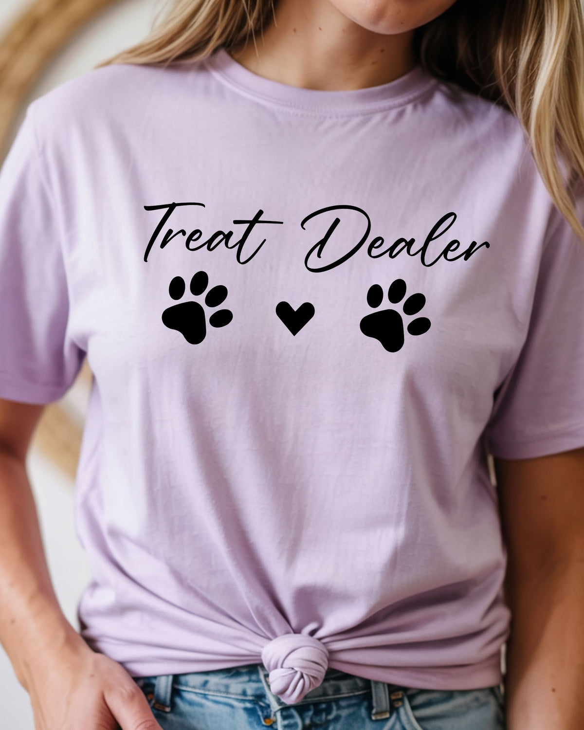 A woman wearing a light lavender t-shirt with a black graphic that reads 'Treat Dealer' with a paw print and heart, paired with blue jeans.