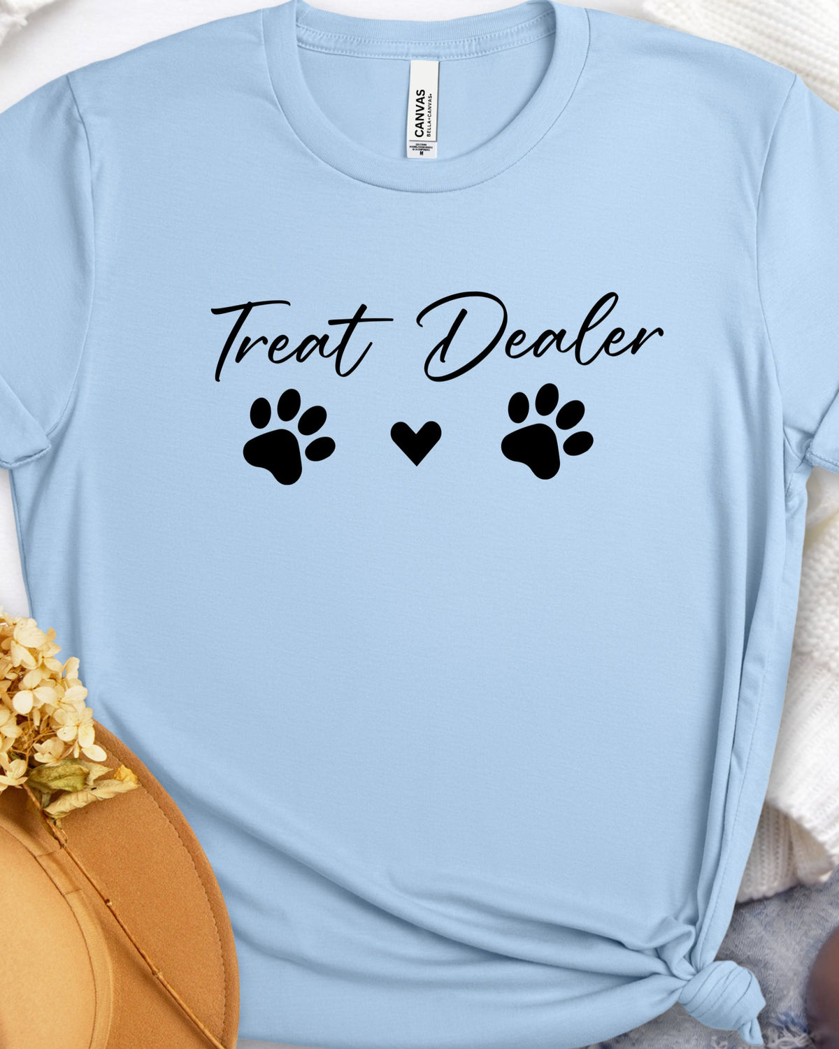 A light blue short-sleeve t-shirt with a black graphic print reading 'Treat Dealer' between two paw prints and a small heart. The shirt is styled flat with a knot at the bottom and dried flowers and a hat in the corners.