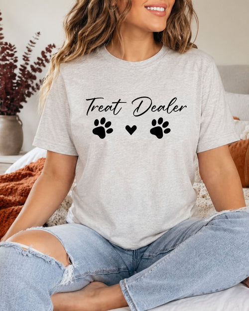 A woman wearing a light heather gray t-shirt with a black graphic that reads 'Treat Dealer' above three paw prints and a heart, paired with light-wash ripped jeans, sitting in a cozy indoor setting.