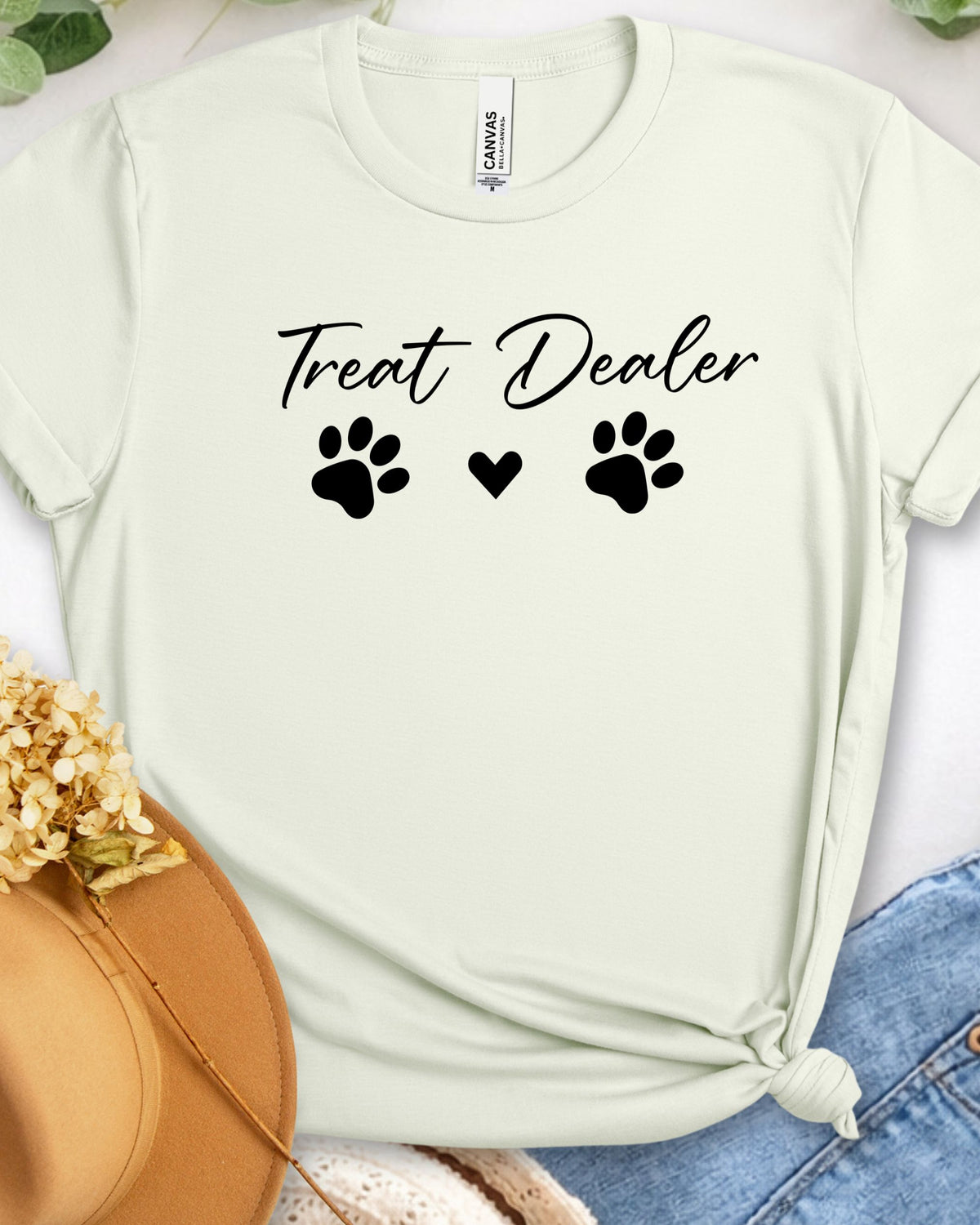 A pale mint green short-sleeve t-shirt with 'Treat Dealer' and paw prints printed in black on the front, styled in a flat lay with a straw hat, blue jeans, and dried flowers.