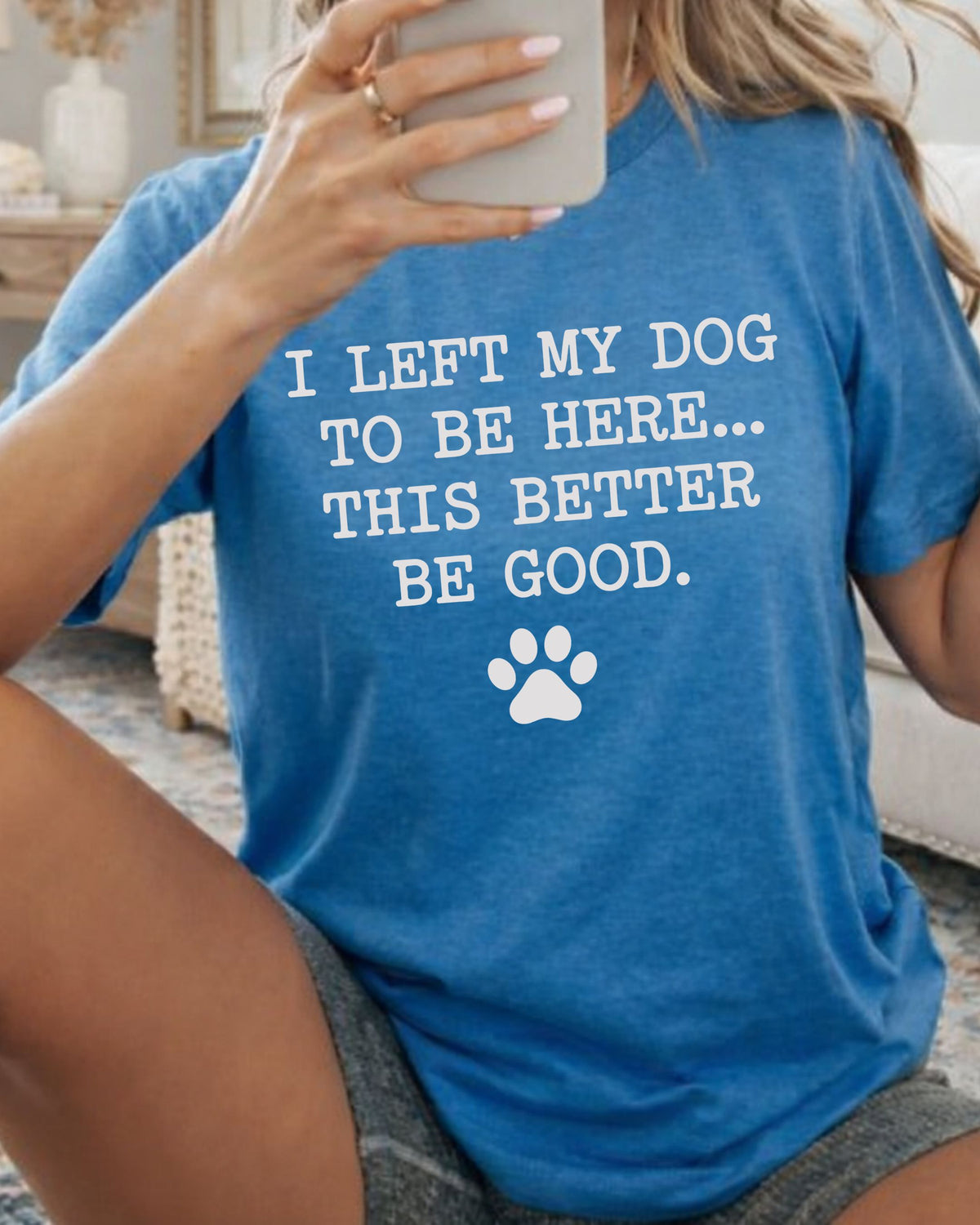 A woman takes a mirror selfie wearing a heather blue graphic t-shirt with the white printed text 'I LEFT MY DOG TO BE HERE.