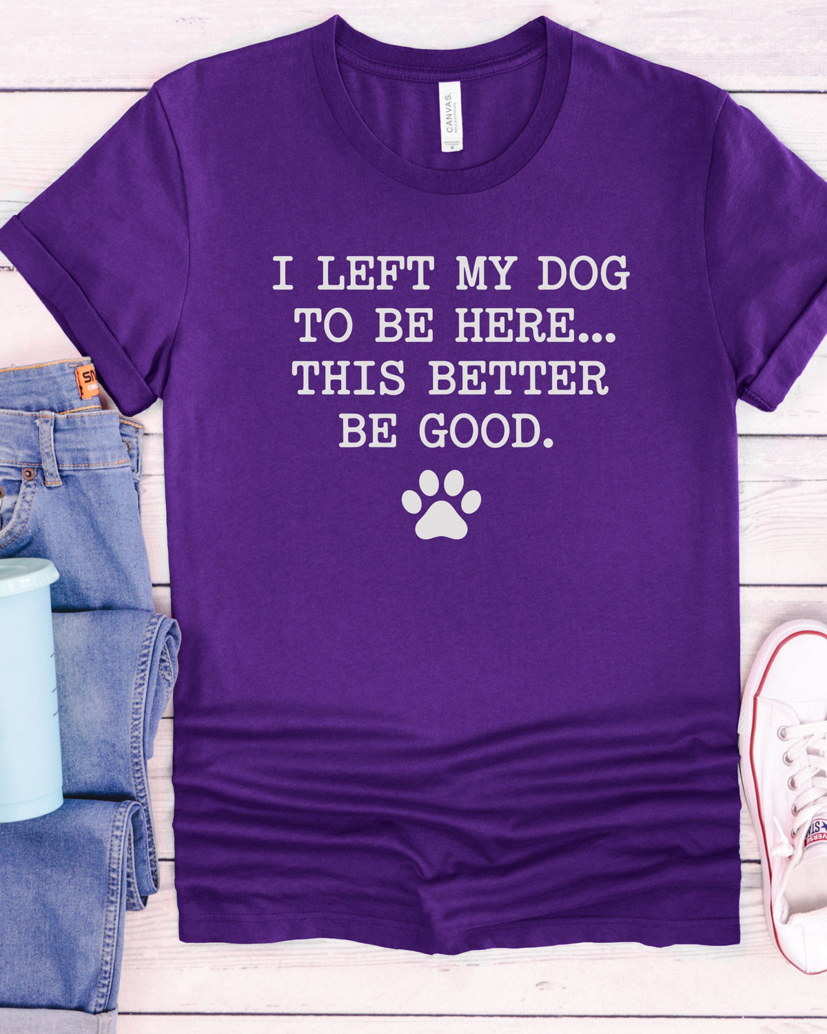 A purple short-sleeve graphic T-shirt laid flat on a light wood background, with white text reading 'I LEFT MY DOG TO BE HERE.