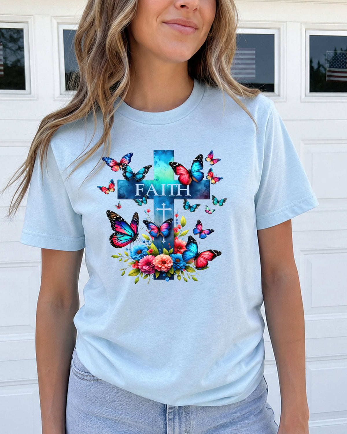 A woman wearing a light blue crewneck t-shirt with a colorful graphic print featuring a cross, butterflies, and flowers with the word 'FAITH' on the front, posed in front of a white garage door.