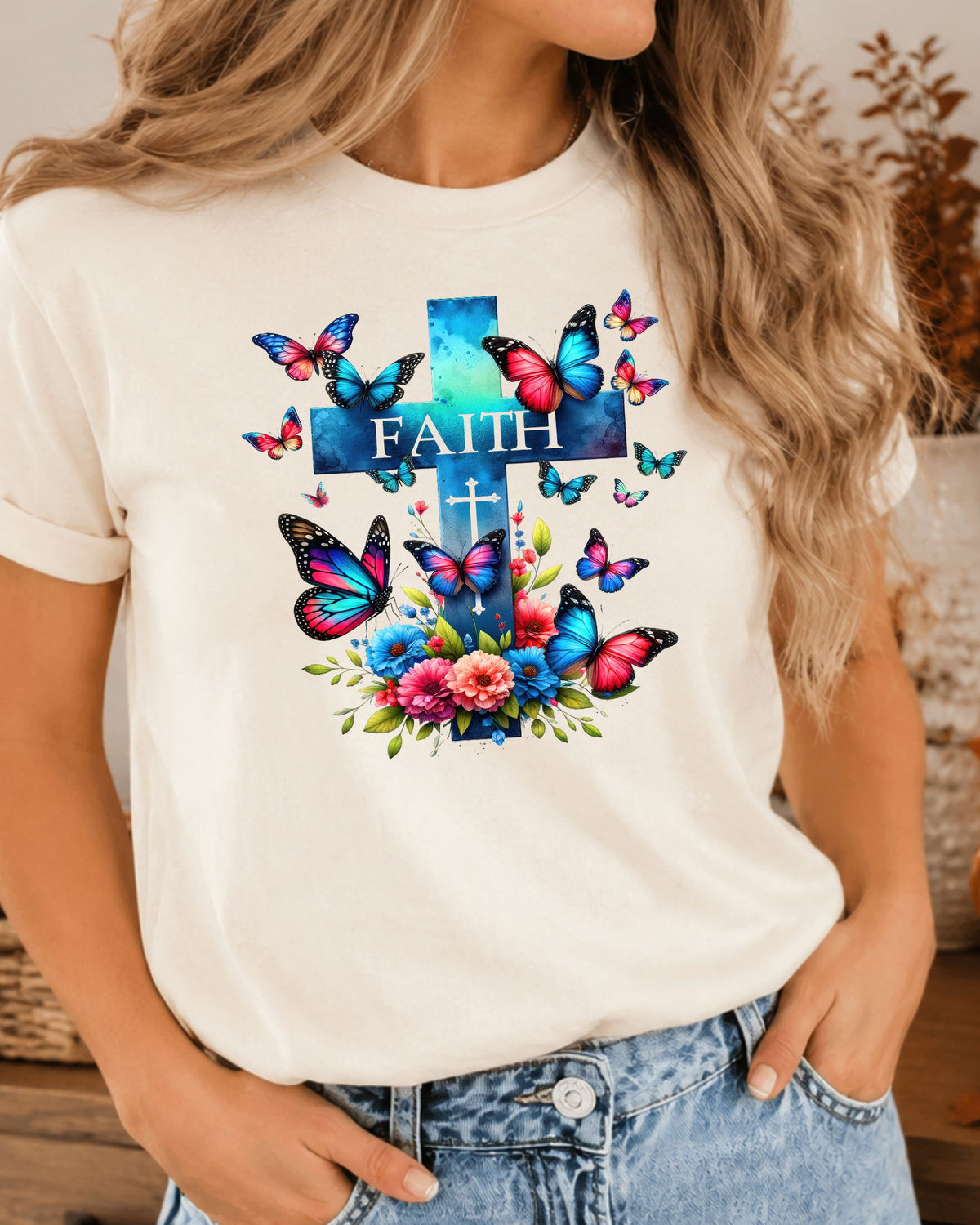 A woman wearing a white short-sleeve t-shirt with a colorful graphic of a cross, butterflies, flowers, and the word 'FAITH' in blue, pink, and purple tones, paired with light-wash denim jeans.