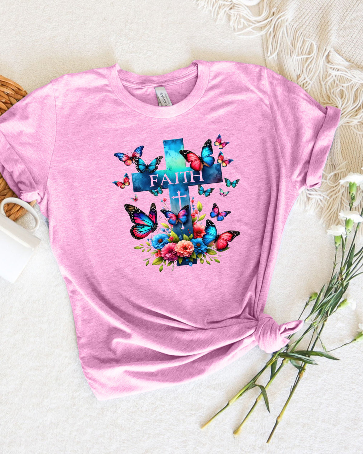 A pink short-sleeve t-shirt with a colorful graphic of a cross, butterflies, and flowers with the word 'FAITH' printed on the chest, styled flat-lay on a white textured blanket with decorative props.
