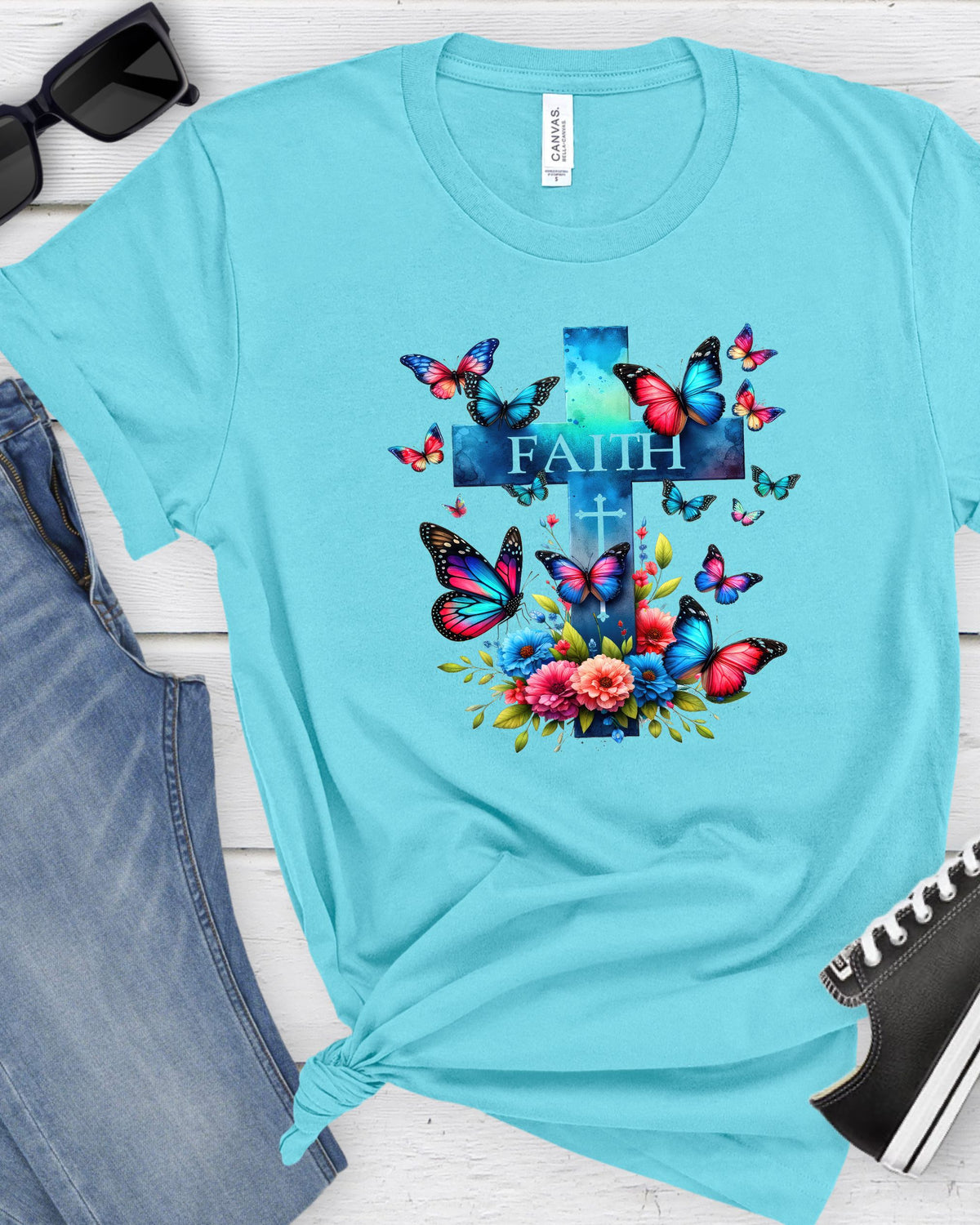 A light blue t-shirt with a colorful graphic of a cross, butterflies, and flowers with the word 'FAITH' printed on the chest, styled in a flat lay with jeans, sunglasses, and a shoe.