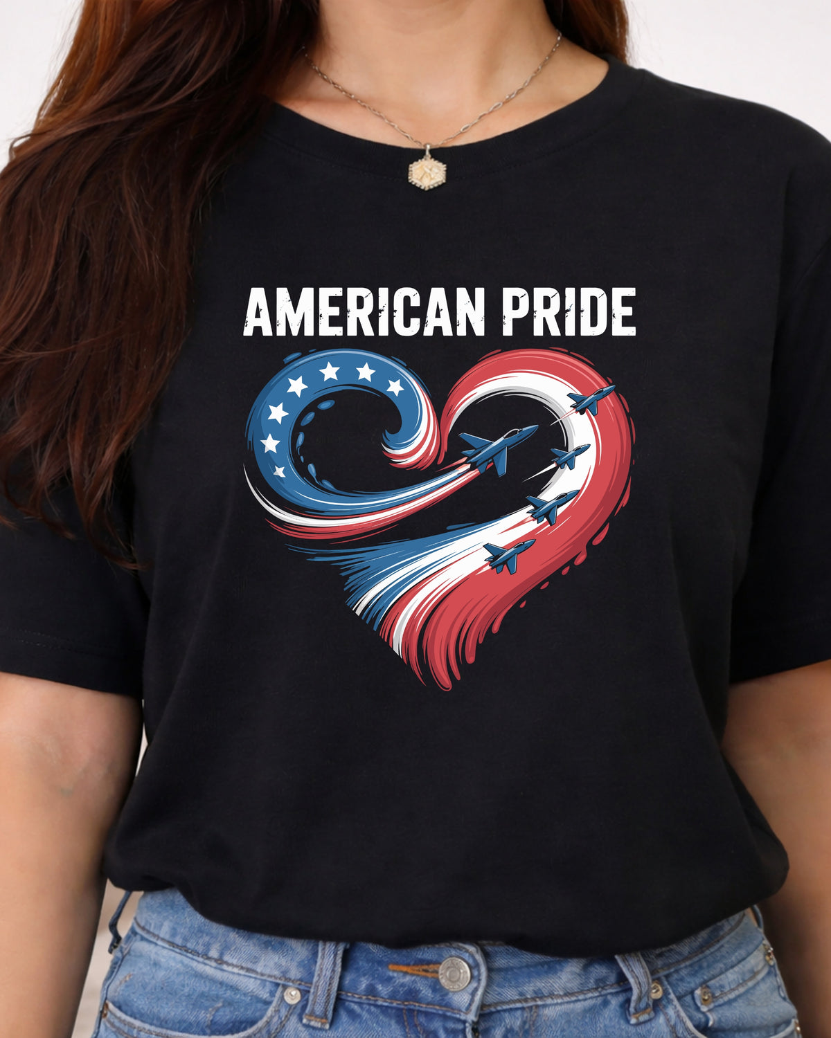 A woman wearing a black short-sleeve t-shirt with a graphic print that reads 'AMERICAN PRIDE' above a heart-shaped swirl of red, white, and blue with fighter jets flying through it, paired with blue jeans.