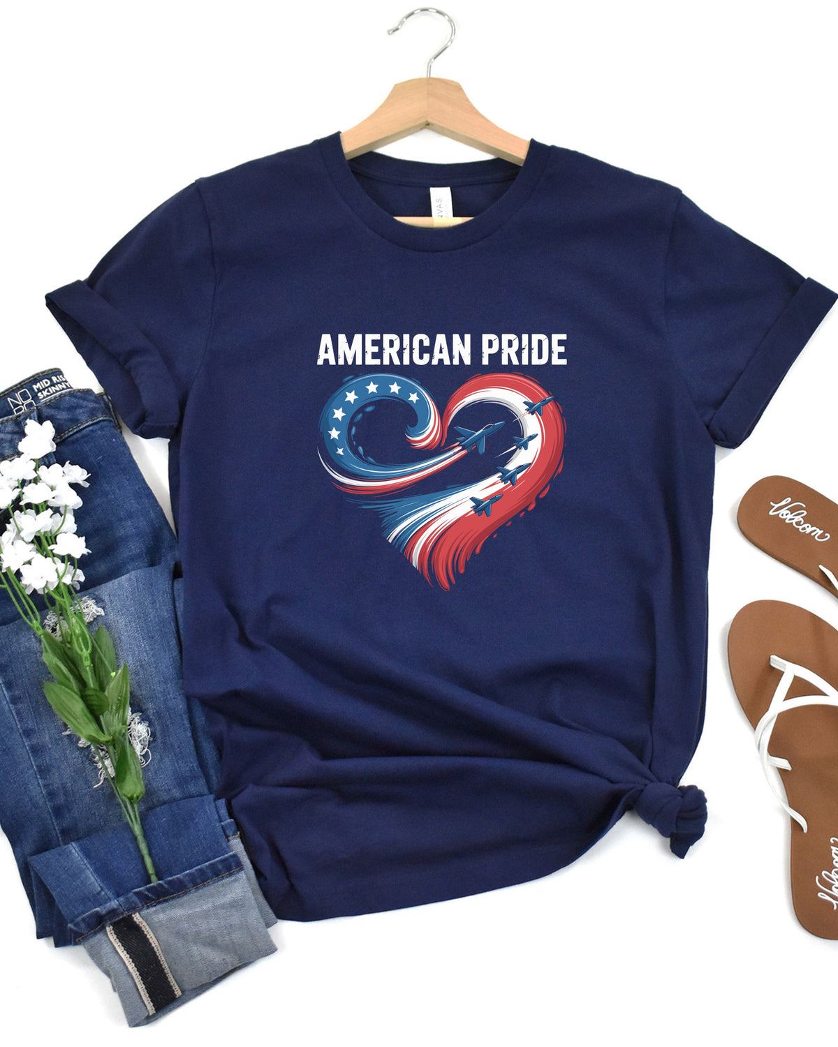 A navy blue t-shirt with a 'AMERICAN PRIDE' graphic on the chest, styled in a flat-lay arrangement with rolled-up jeans, white flowers, and brown sandals on a white background.