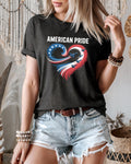 A woman wearing a dark charcoal heather t-shirt with a patriotic graphic of a heart made of stars and stripes, paired with distressed denim shorts in a bohemian-style room.