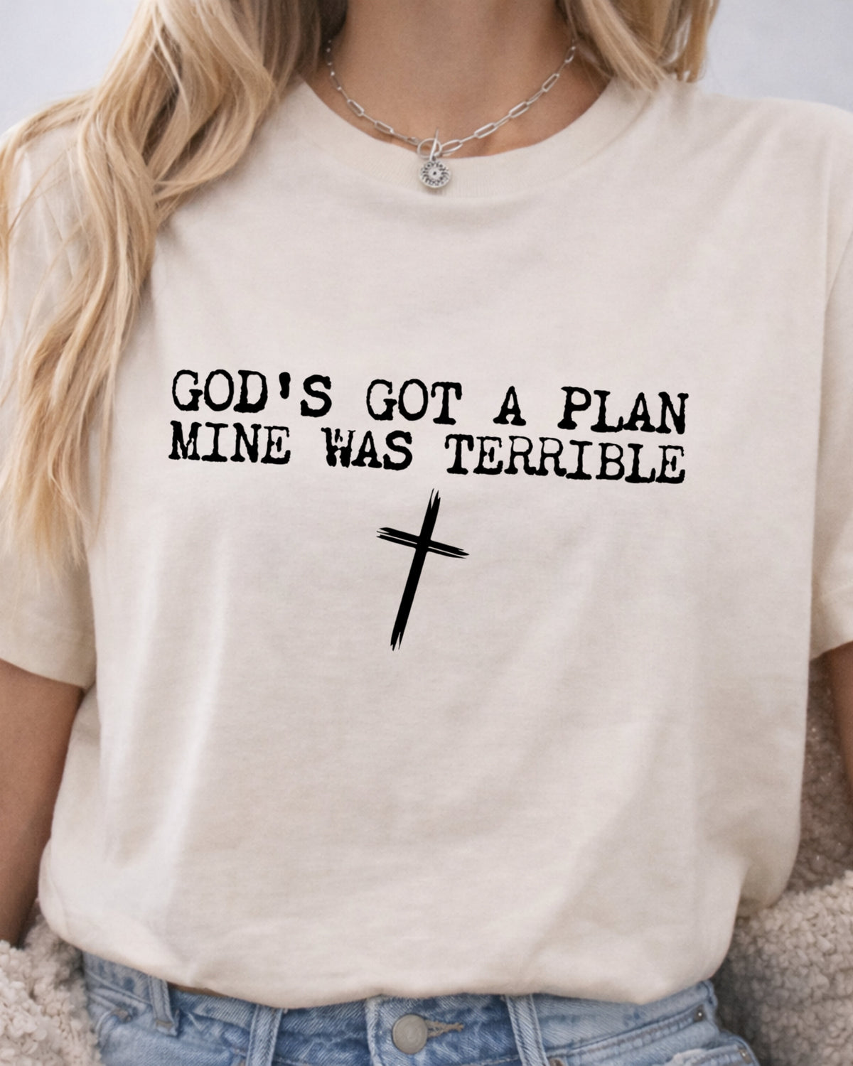 A woman wearing a cream-colored graphic t-shirt with the text 'GOD'S GOT A PLAN MINE WAS TERRIBLE' and a black cross printed on the front, paired with blue jeans.
