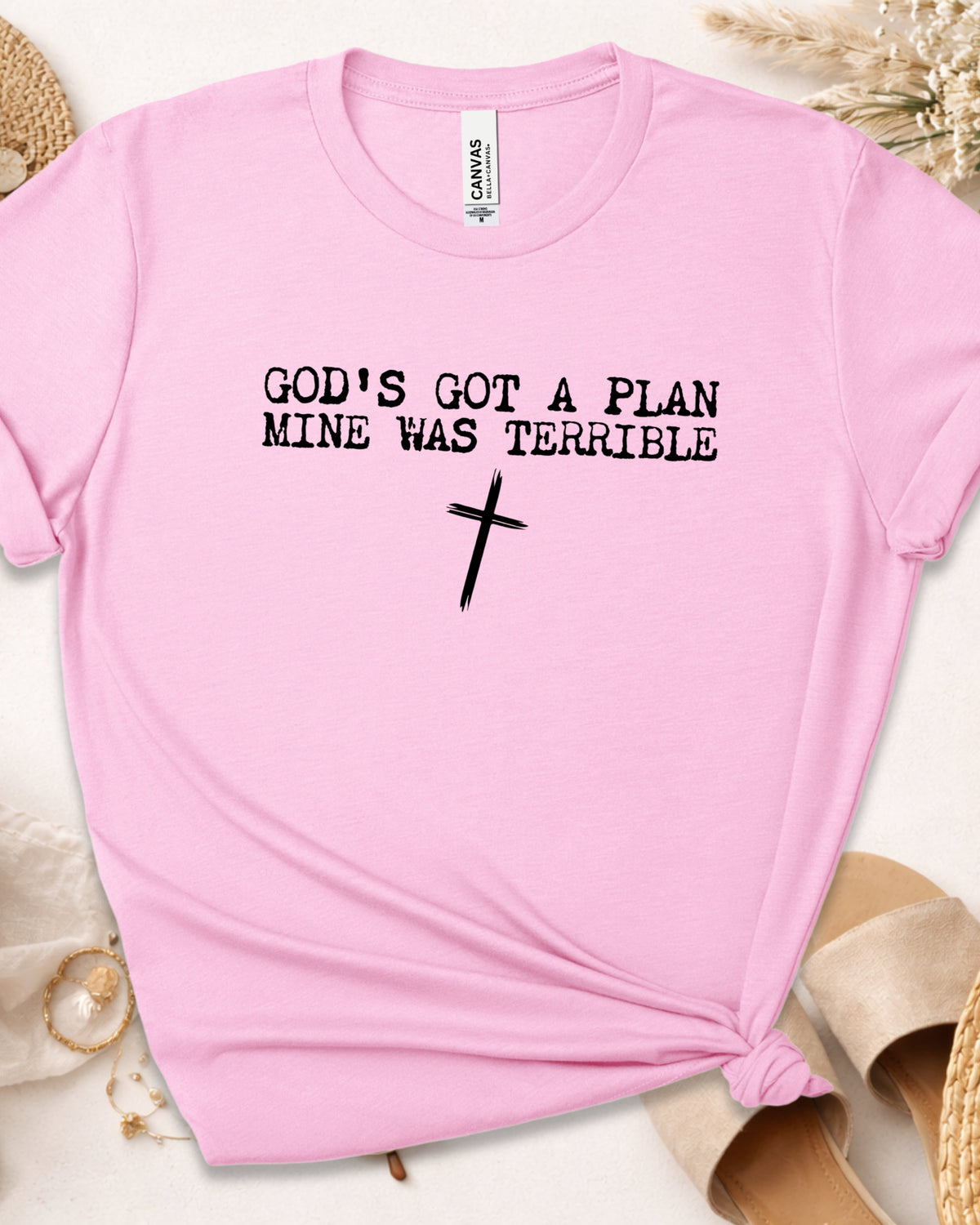 A light pink short-sleeve t-shirt with the black printed text 'GOD'S GOT A PLAN MINE WAS TERRIBLE' and a black cross graphic, styled in a flat lay with beige sandals and dried grass.