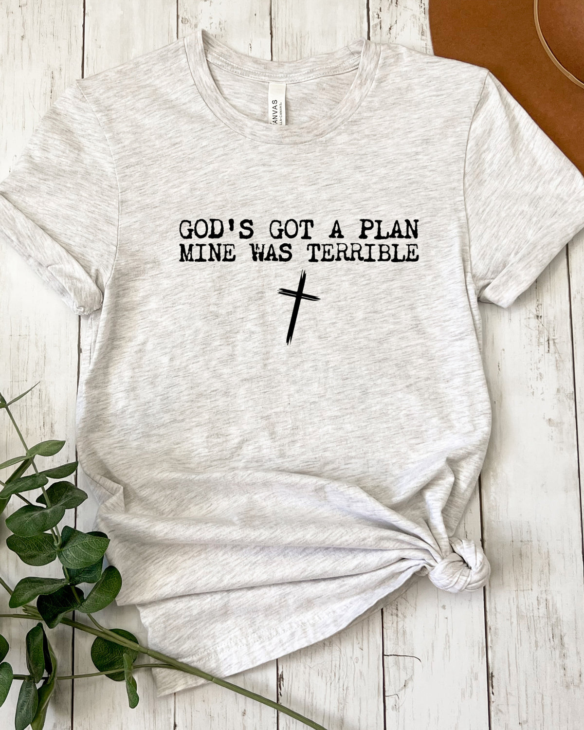 A light heather gray t-shirt with black text reading 'GOD'S GOT A PLAN MINE WAS TERRIBLE' and a black cross graphic, styled flat on a white wood plank background with a green plant and a brown hat visible as props.