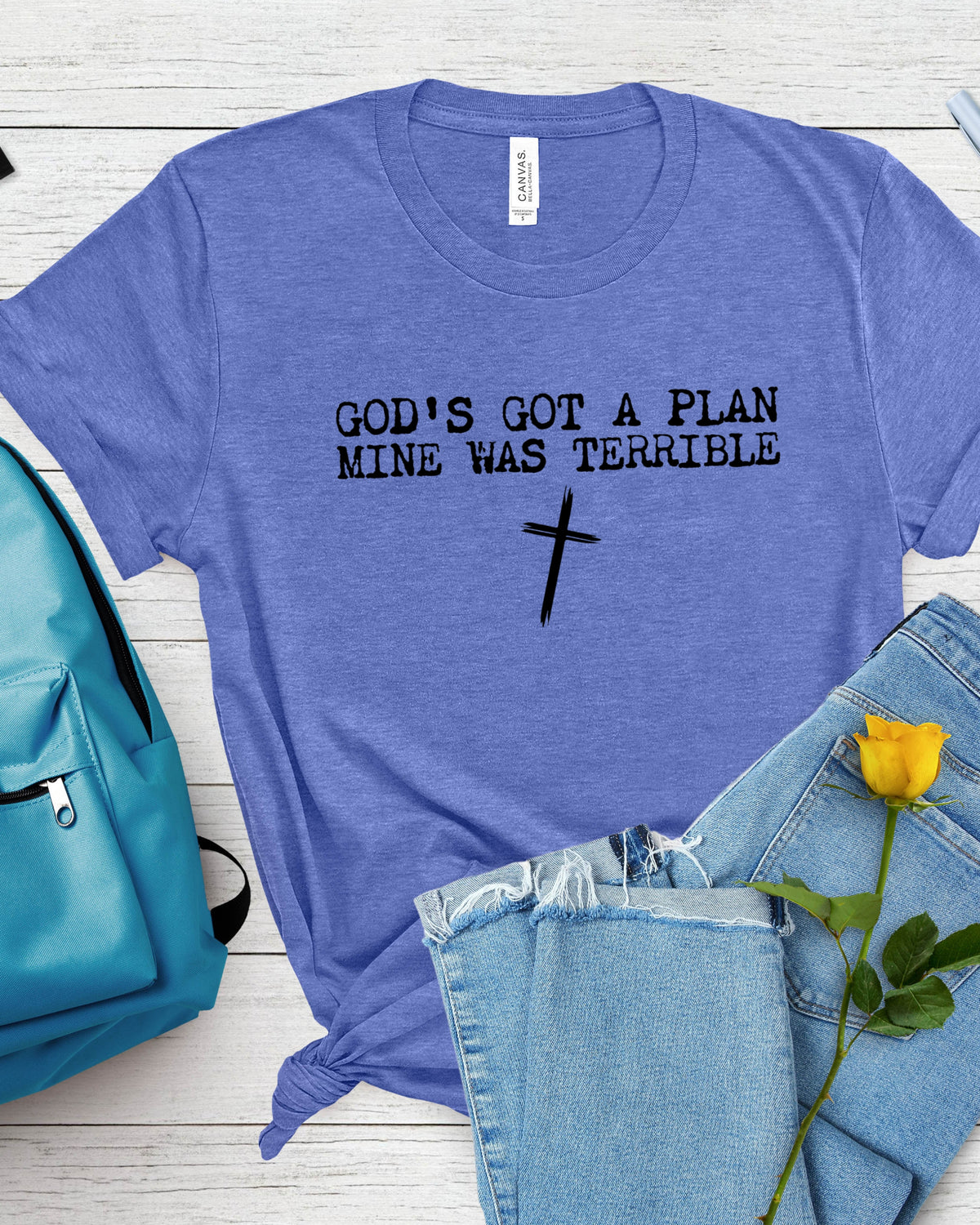 A heathered periwinkle blue t-shirt with the black text 'GOD'S GOT A PLAN MINE WAS TERRIBLE' and a black cross graphic, styled in a flat lay with light-wash jeans, a yellow rose, and a teal backpack on a white wood background.