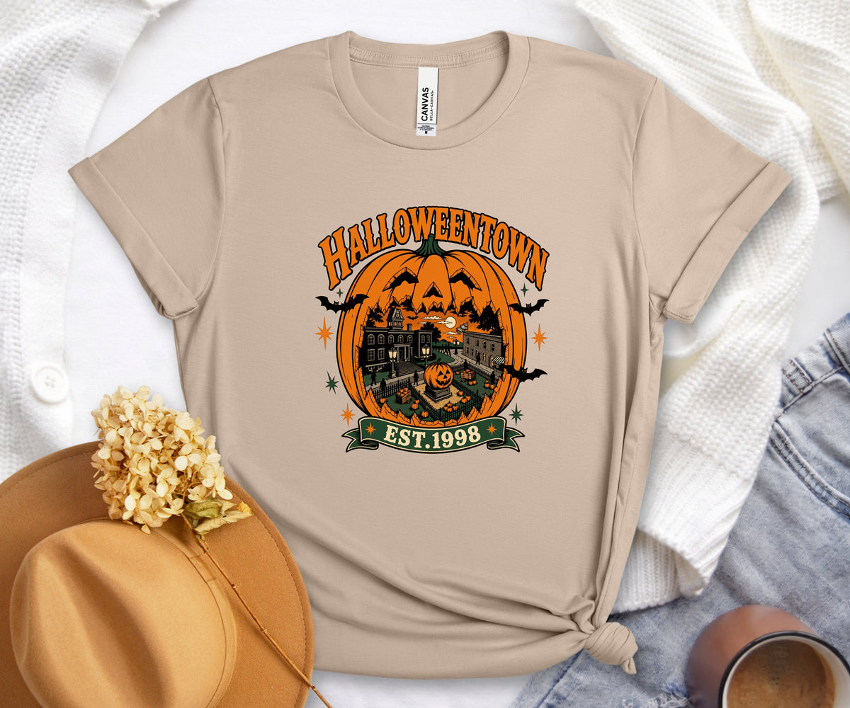 A beige graphic t-shirt featuring a Halloween-themed design with a jack-o'-lantern, haunted house, bats, and the text 'HALLOWEENTOWN EST. 1998'.