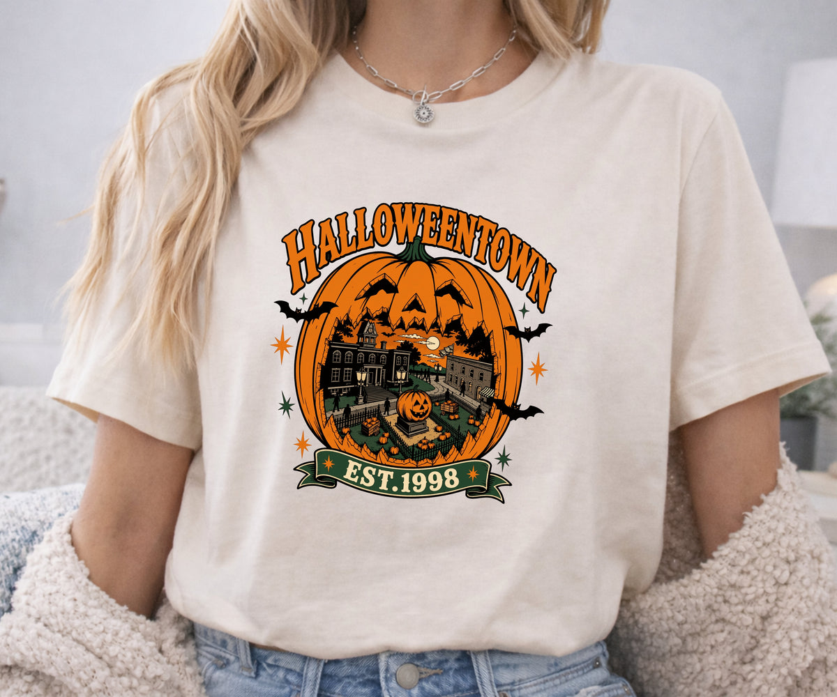 A woman wearing a cream-colored graphic t-shirt with a Halloween-themed design that reads 'HALLOWEENOWN EST. 1998' featuring an orange jack-o'-lantern, haunted house, and bats.