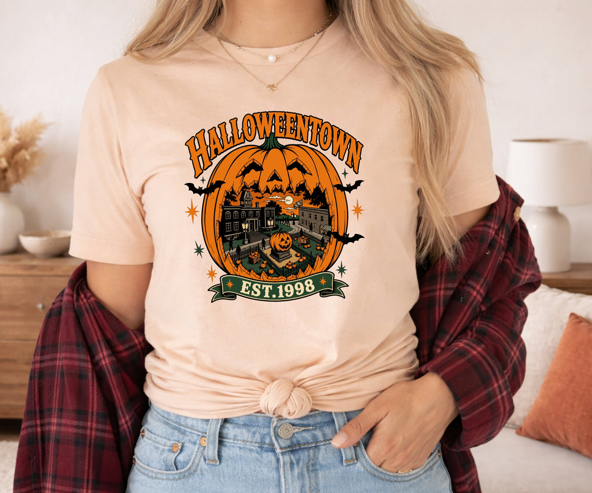 A woman wearing a light beige graphic t-shirt with a Halloween-themed 'Halloweentown Est. 1998' print, styled with a red plaid flannel shirt tied around her shoulders and light blue jeans.