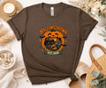 A brown short-sleeve graphic T-shirt laid flat on a light surface with seasonal props. The shirt has a large Halloween-themed front print reading 'HALLOWEENTOWN EST.