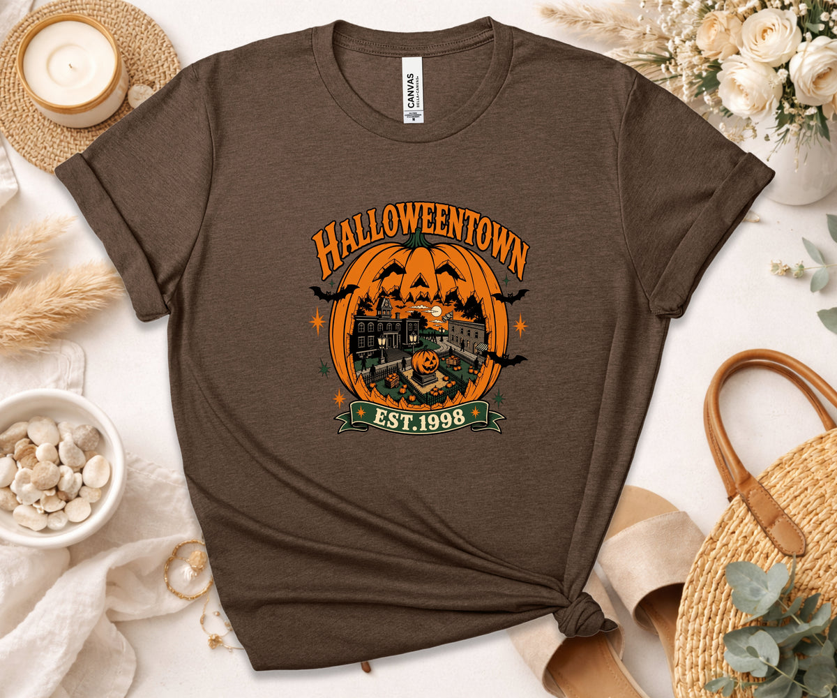 A brown short-sleeve graphic T-shirt laid flat on a light surface with seasonal props. The shirt has a large Halloween-themed front print reading 'HALLOWEENTOWN EST.