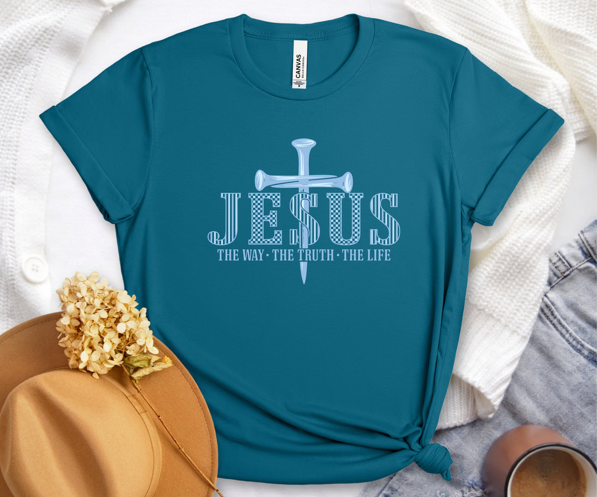 A teal short-sleeve t-shirt with a white graphic print of a nail-shaped cross and the text 'JESUS THE WAY - THE TRUTH - THE LIFE'. The shirt is styled in a flat-lay arrangement with a tan hat, dried flowers, a white knit cardigan, and denim jeans.