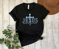 A black short-sleeve t-shirt with a light blue graphic reading 'JESUS' and 'THE WAY - THE TRUTH - THE LIFE' beneath a cross, styled in a flat lay on a whitewashed wood background with a brown hat and green leaves.