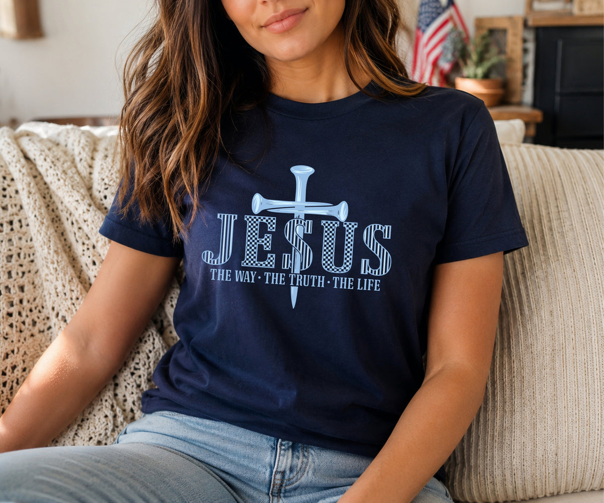A woman wearing a navy blue graphic t-shirt with a light blue cross and the text 'JESUS THE WAY - THE TRUTH - THE LIFE' sitting on a beige couch.