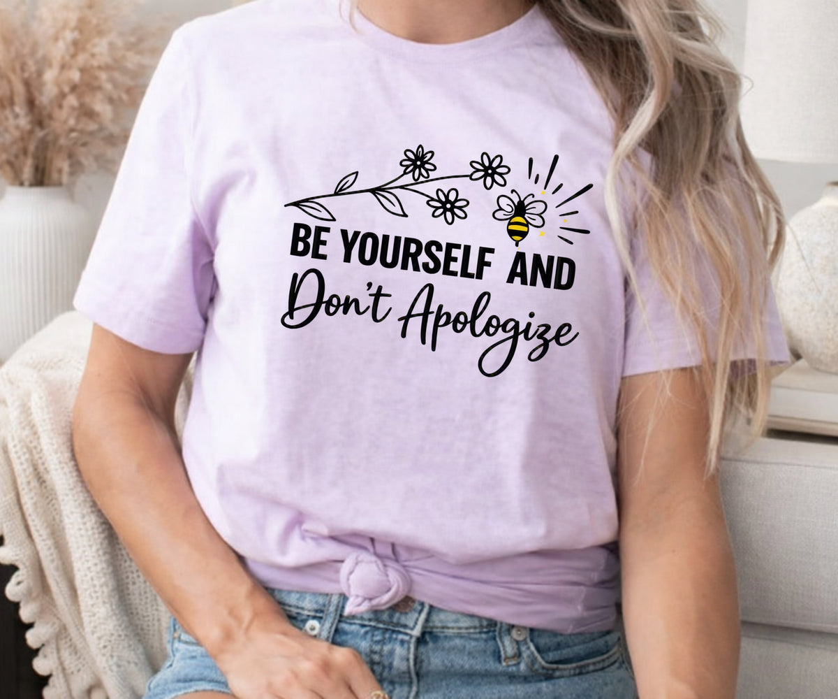 A woman wearing a light lavender graphic t-shirt with the text 'BE YOURSELF AND Don't Apologize' in black script and floral/bee illustrations. The t-shirt is styled with a knot at the bottom hem, paired with blue denim shorts in a cozy indoor setting.