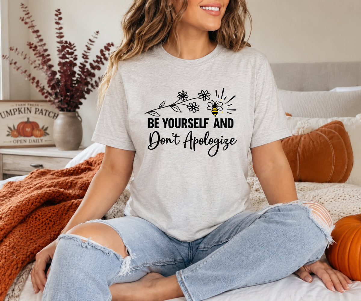 A woman sitting cross-legged on a bed wearing a light heather gray graphic t-shirt with a black text print that reads 'BE YOURSELF AND Don't Apologize' with a bee and floral design. She is wearing distressed light blue jeans in a cozy bedroom setting with autumn decor.