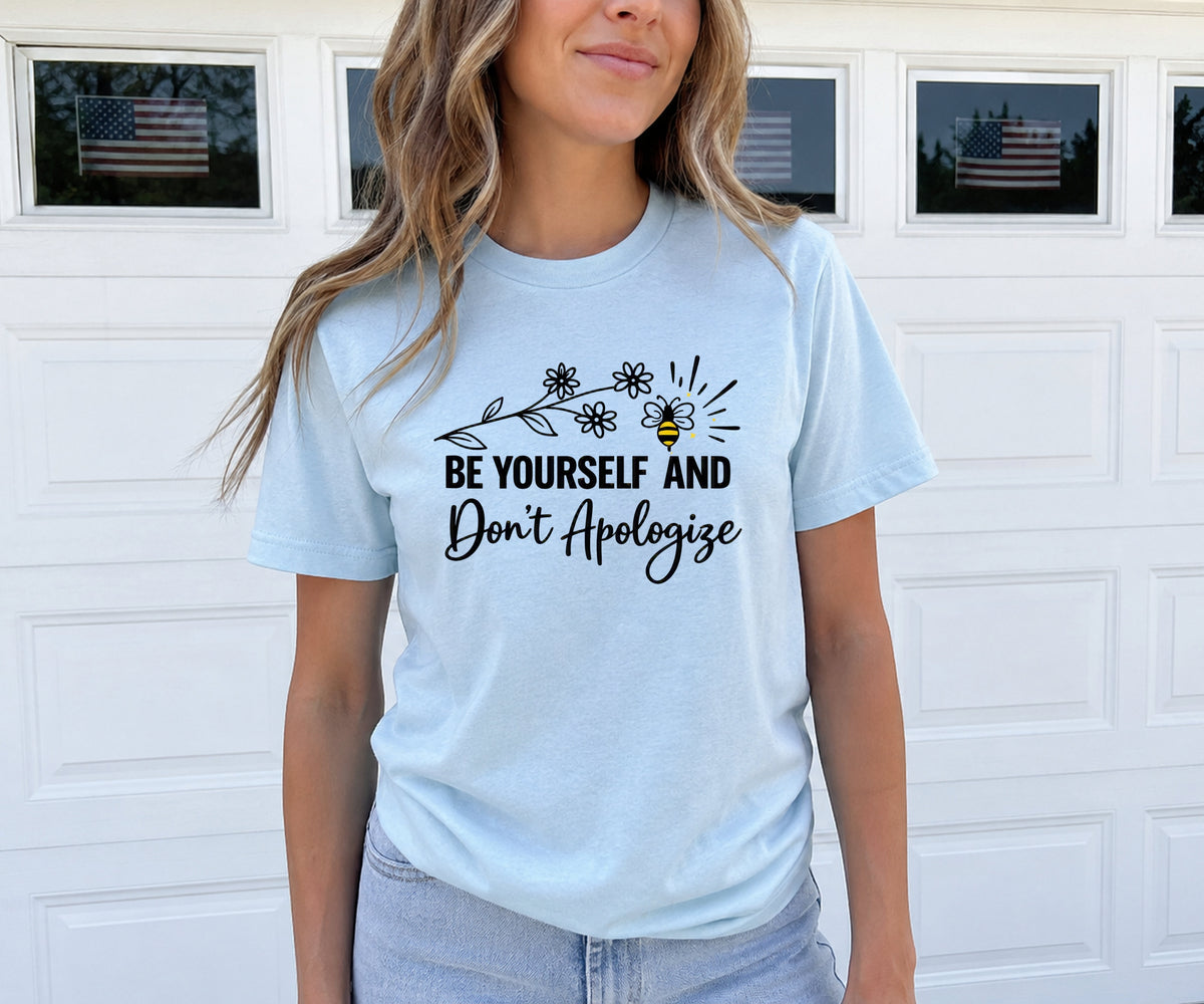 A woman wearing a light blue graphic t-shirt with the text 'BE YOURSELF AND Don't Apologize' and a bee illustration, standing in front of a white garage door.