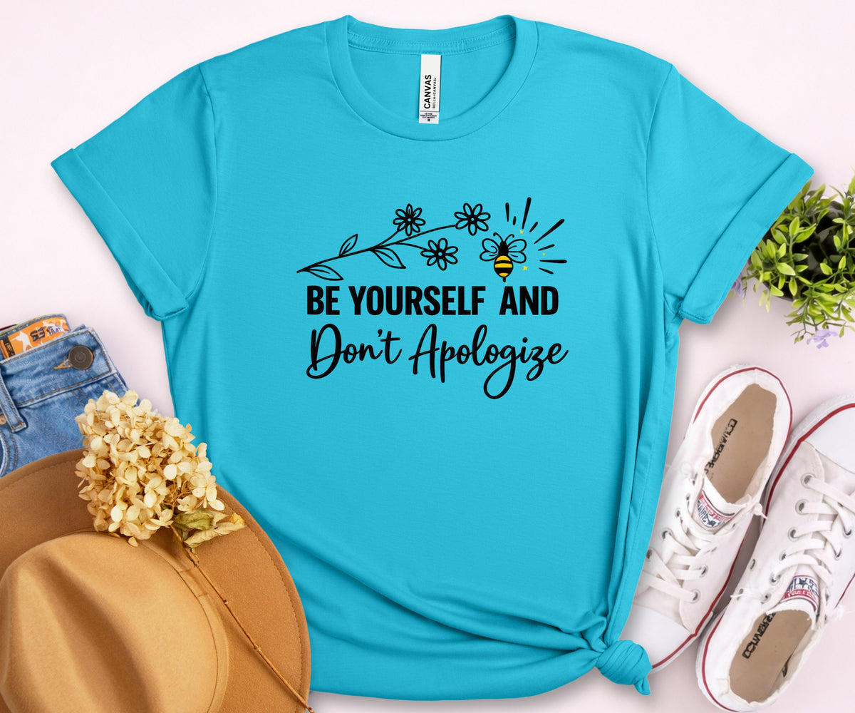 A bright turquoise short-sleeve graphic T-shirt laid flat on a pale pink background, styled with a tan hat, dried flowers, blue jeans, white sneakers, and a small green plant. The shirt has a black floral branch and bee graphic above the phrase “BE YOURSELF AND Don’t Apologize.
